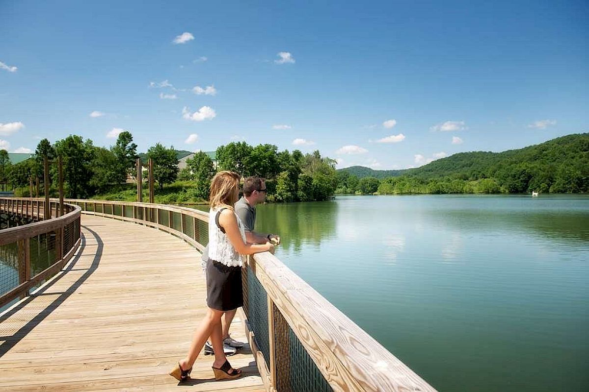 A couple stands on a wooden boardwalk overlooking a tranquil lake, surrounded by greenery and hills under a clear blue sky.