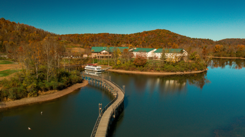 A lake with a winding boardwalk leading to a docked boat, surrounded by buildings and autumn trees, under a clear blue sky.