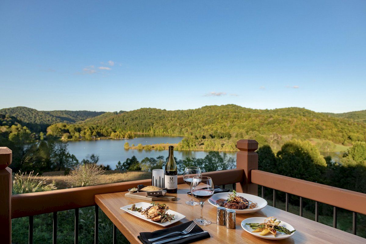 A table with plates of food and wine overlooks a scenic view of a lake and green hills under a clear blue sky.