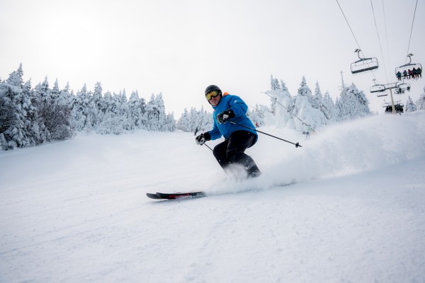 A skier in a blue jacket carves through fresh powder on a snowy slope, trees lined with frost and a chairlift in the background.