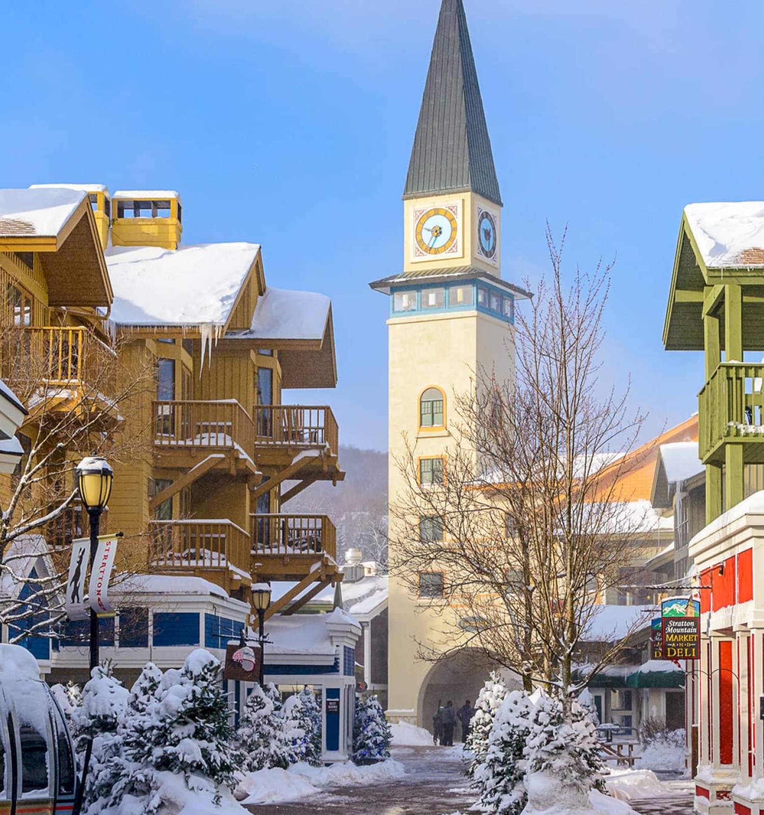 A snowy street scene in a quaint town, with colorful buildings, a clock tower in the distance, and a light dusting on the roofs.