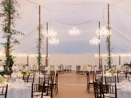 An elegant beachside wedding setup with string lights, chandeliers, and neatly arranged round tables under a tent, wooden chairs, and a sandy floor.
