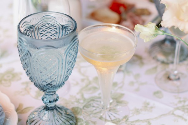 A fancy table setting with pastel flowers, glassware, and a small plate of elegant finger foods on a lace-patterned tablecloth, inviting guests.