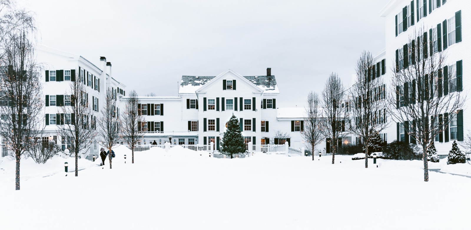 A snow-covered campus blanketed in white, with leafless trees lining walkways, pale buildings, and a quiet, serene winter scene.