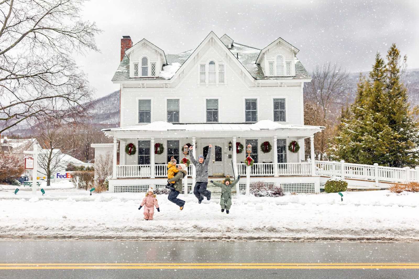 A family joyfully jumps in front of a festive, snowy house decorated with wreaths during winter's snowfall.