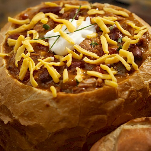 A bread bowl filled with chili, topped with shredded cheese and a dollop of sour cream, served in a rustic crusty loaf.