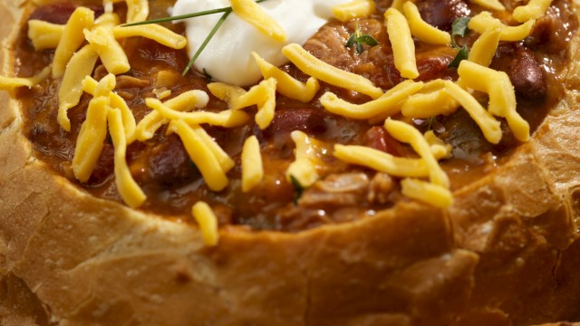 A bread bowl filled with chili, topped with shredded cheese and a dollop of sour cream, served in a rustic crusty loaf.