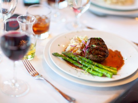 A plated steak with green asparagus, mashed potatoes, and red wine sauce on a fine dining table, surrounded by wine glasses and cutlery.